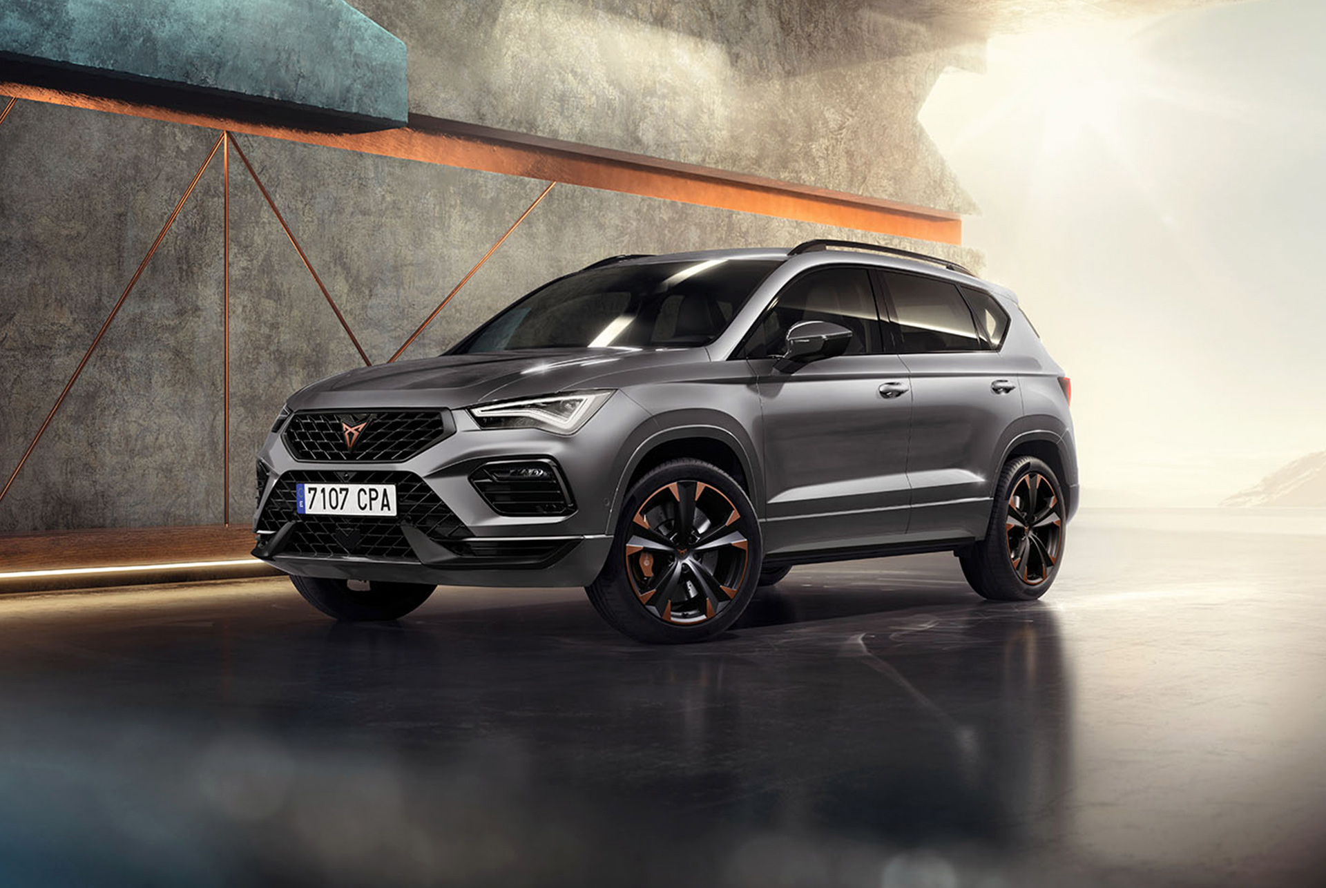 Grafiitinharmaa 2024 CUPRA Ateca, urheilullinen ja kompakti SUV pysäköitynä modernin teollisuusrakennuksen eteen. Muotoilussa korostuvat aerodynaamiset linjat, led-ajovalot ja omaleimaiset kevytmetallivanteet kuparisin somistein. Taustalla geometrinen seinäkuvio, joka tuo esiin auton sulavalinjaisen muotokielen.